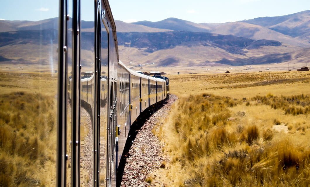 A train tour moves along a curved track through a dry, grassy landscape with mountains in the distance, possibly in Peru or Bolivia. The train’s reflection is visible on its shiny side in bright daylight.