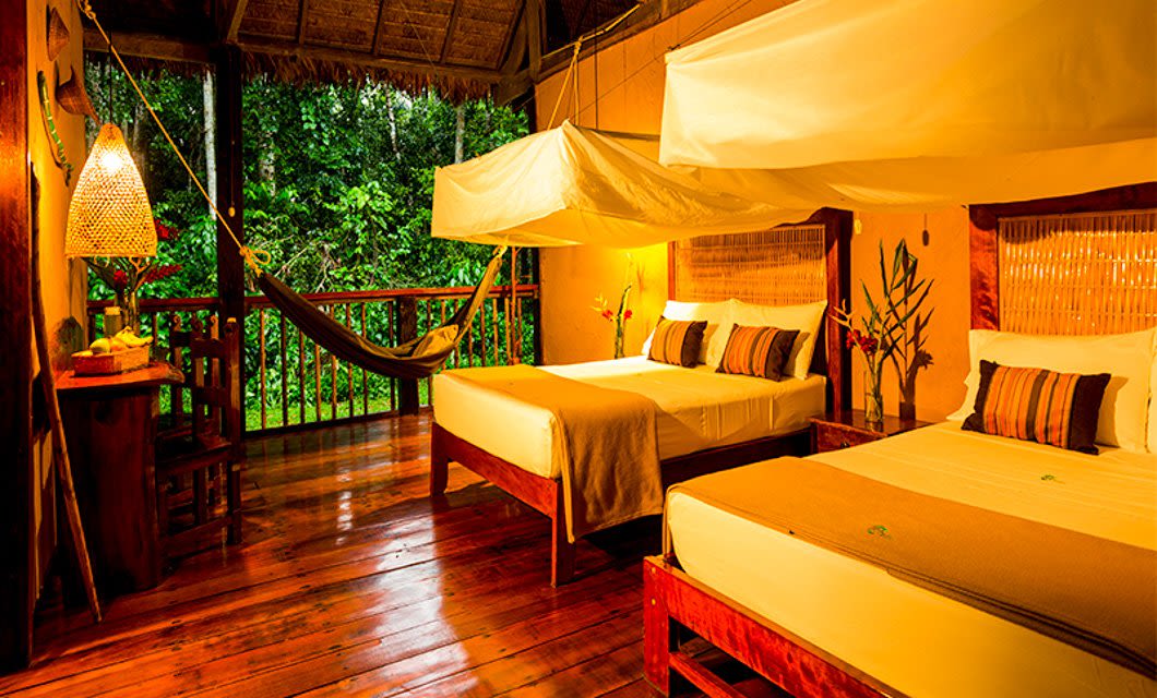 A cozy, warmly lit tropical cabin at Tambopata Research Center with two canopy-covered beds, a hammock on the wooden balcony, a desk with a lamp, and lush green jungle visible outside through open walls.