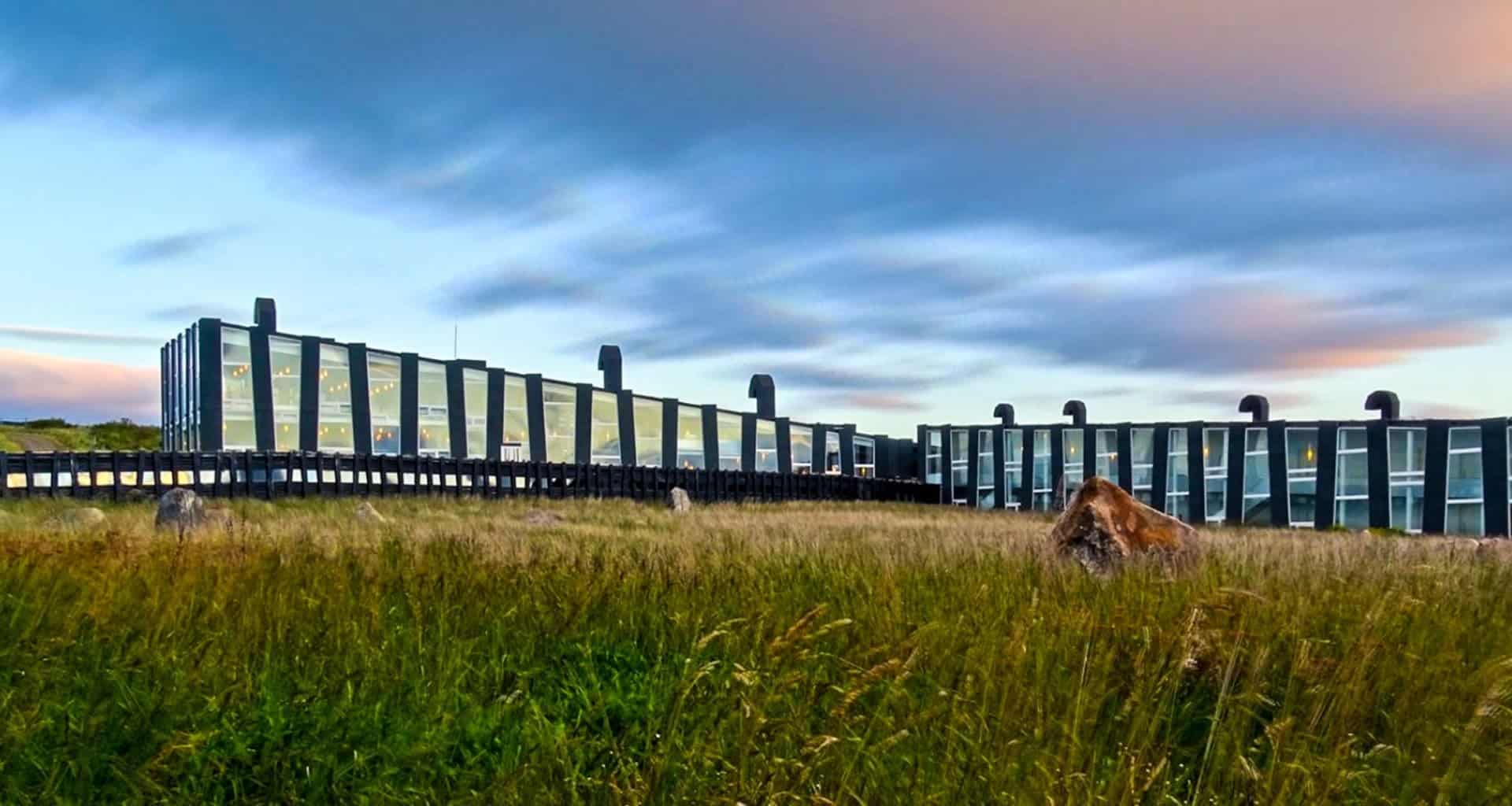 Modern building with large glass windows and dark-colored vertical columns, reminiscent of Remota Patagonia, set in a grassy field under a dramatic, cloudy sky at sunset.