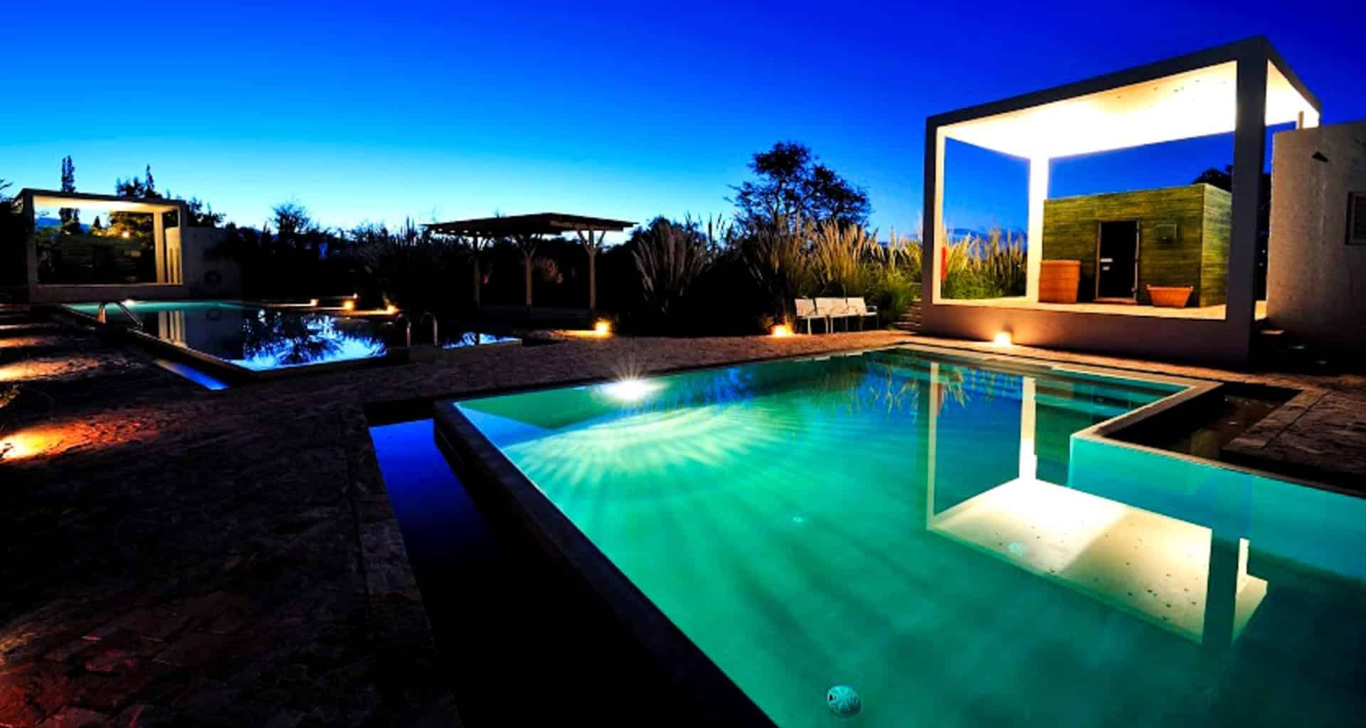 A modern swimming pool at dusk, illuminated with underwater lights, surrounded by contemporary cabanas and lush landscaping at the luxury hotel Explora Atacama, with a deep blue evening sky above the breathtaking Atacama Desert.
