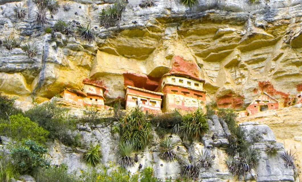 Colorful ancient cliffside tombs built into a rocky canyon wall near Chachapoyas, surrounded by green vegetation and distinctive yellow and orange stone layers.