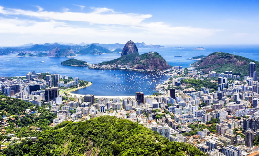 Aerial view of Rio de Janeiro with Sugarloaf Mountain, Guanabara Bay, and city buildings surrounded by lush green hills under a clear blue sky, capturing the vibrant beauty of this iconic tropical island city.