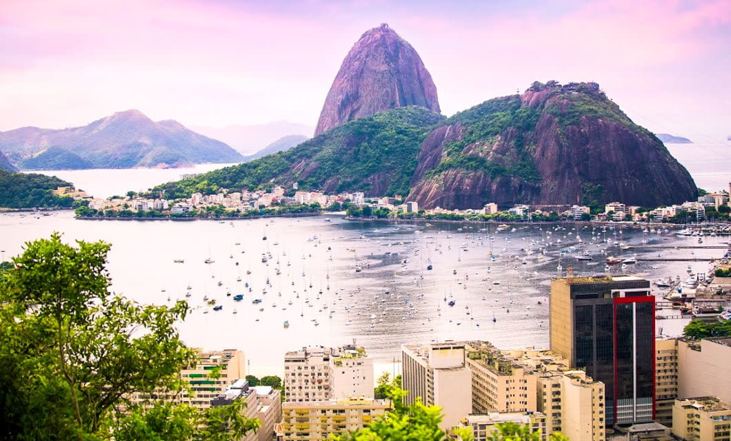 A scenic view of Rio de Janeiro along the historic Route of Gold, with Sugarloaf Mountain rising in the background, a bay filled with boats, and city buildings in the foreground under a pink and purple sky.