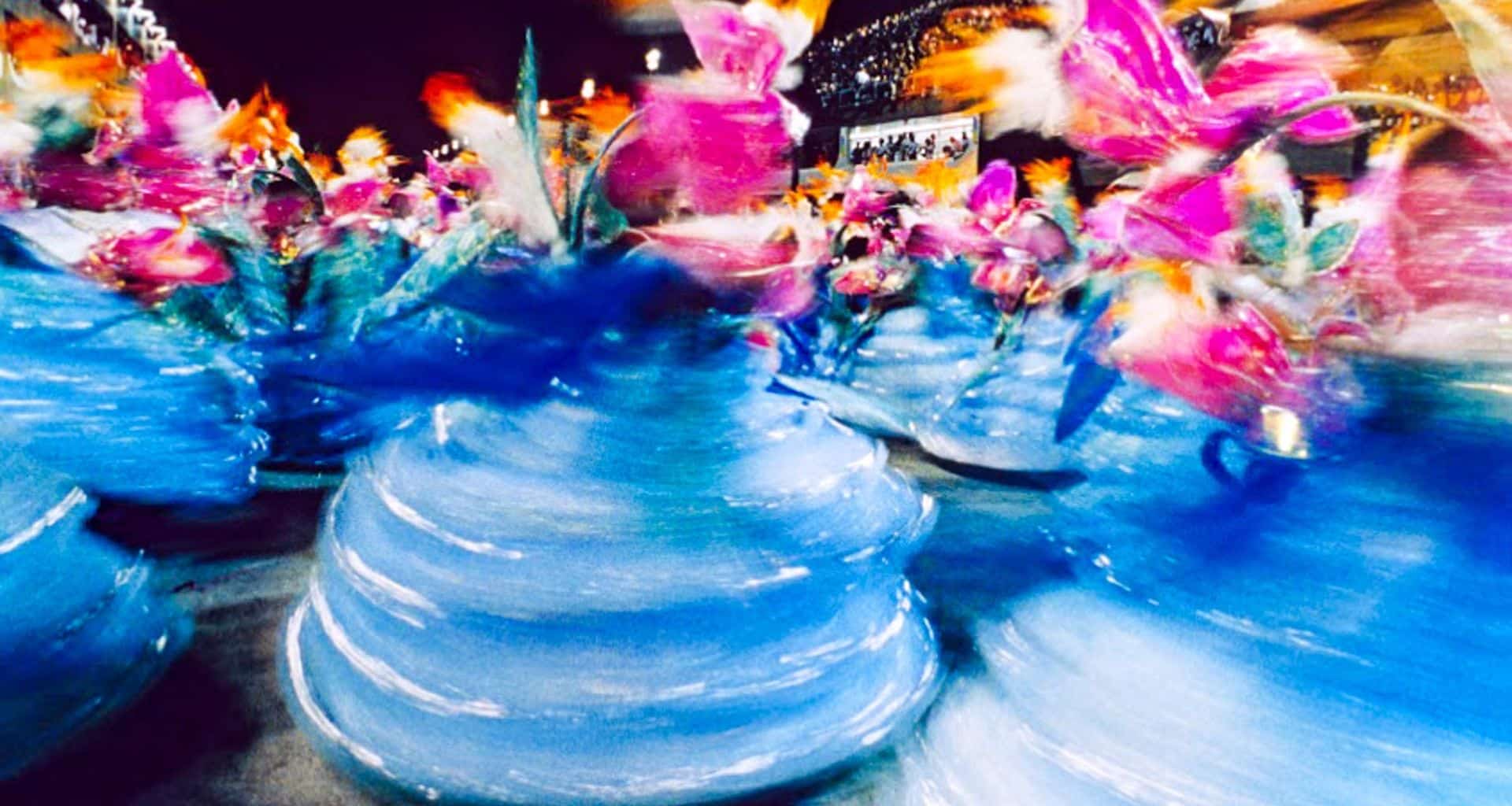 Blurred image of dancers in swirling blue and pink costumes, likely at Rio's Carnival or a lively parade, with vibrant colors and motion creating a sense of energy and celebration—a highlight for those drawn to Rio tourism.