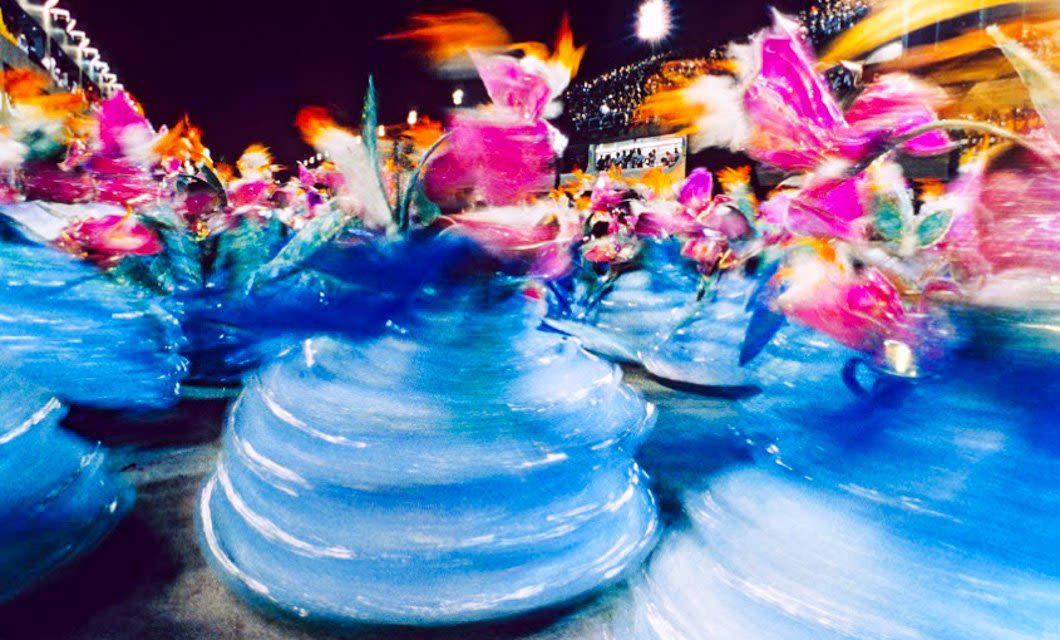 Dancers in vibrant blue swirling costumes and colorful headpieces move energetically during Rio’s Carnival parade, creating a dynamic and blurred effect that captures the excitement of the celebration.