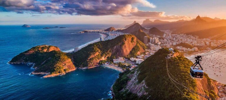 A cable car ascends over Sugarloaf Mountain, offering a panoramic view of Rio de Janeiro on Brazil Tours. The city, coastline, and distant mountains glow in golden sunlight beneath a partly cloudy sky.