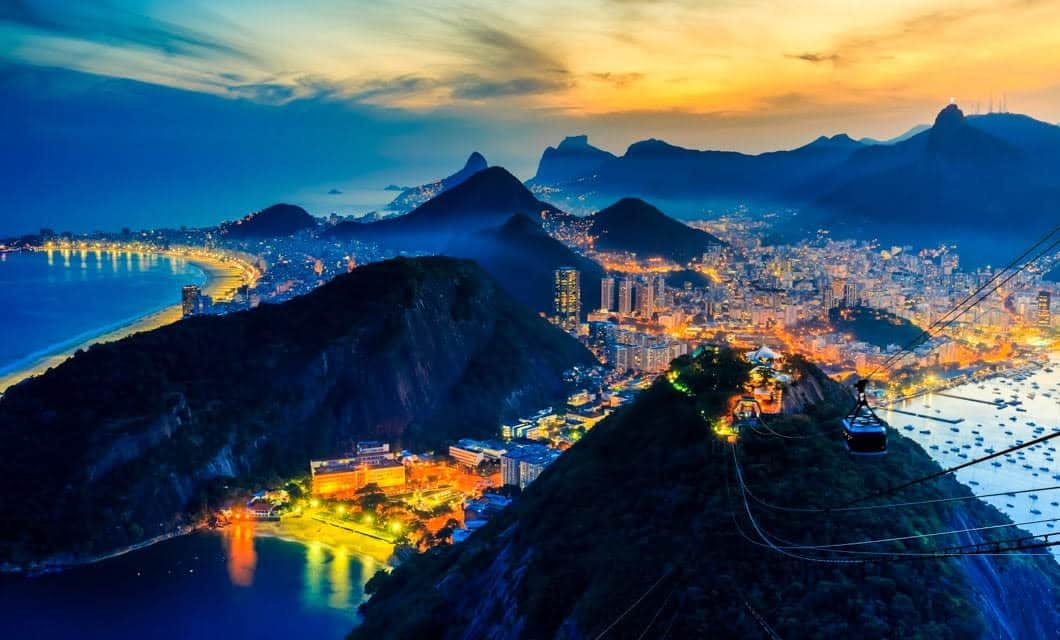 A vibrant, colorful sunset view over Rio de Janeiro, Brazil, showing city lights, mountains, the coastline, and Sugarloaf Mountain with cable cars in the foreground—echoing the adventurous spirit of Incas exploring new horizons.