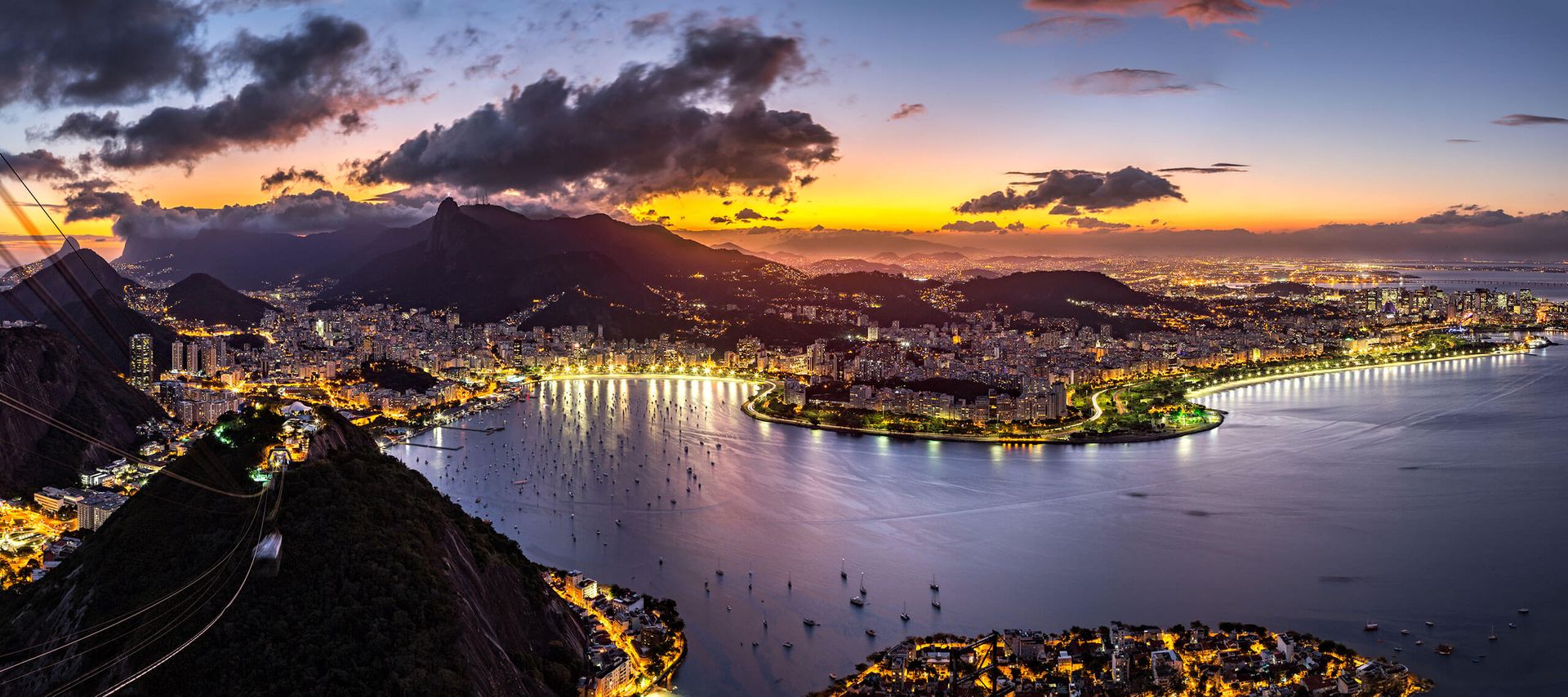 A panoramic aerial view of Rio de Janeiro at sunset, one of South America's Cultural Capitals, showing city lights, mountains, and the curved coastline with boats anchored in the bay under a colorful sky.