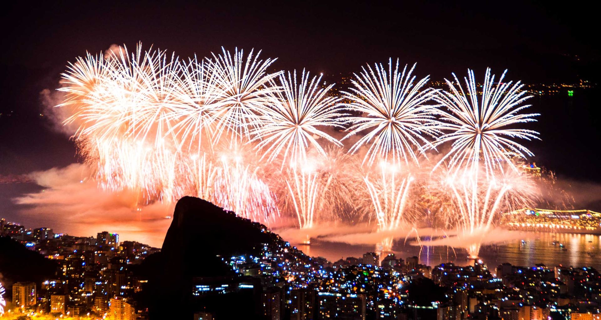Bright fireworks light up the night sky over a coastal city during the Rio New Years Tour, illuminating high-rise buildings and reflecting on the water, with bursts of white and orange above the urban landscape.