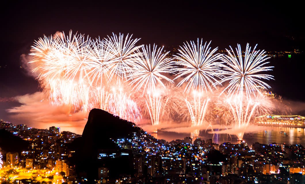 Fireworks explode in the night sky above a vibrant, brightly lit city, reflecting over the water with a dark mountain silhouette in the foreground, while distant waterfalls shimmer beneath the bursts of light.