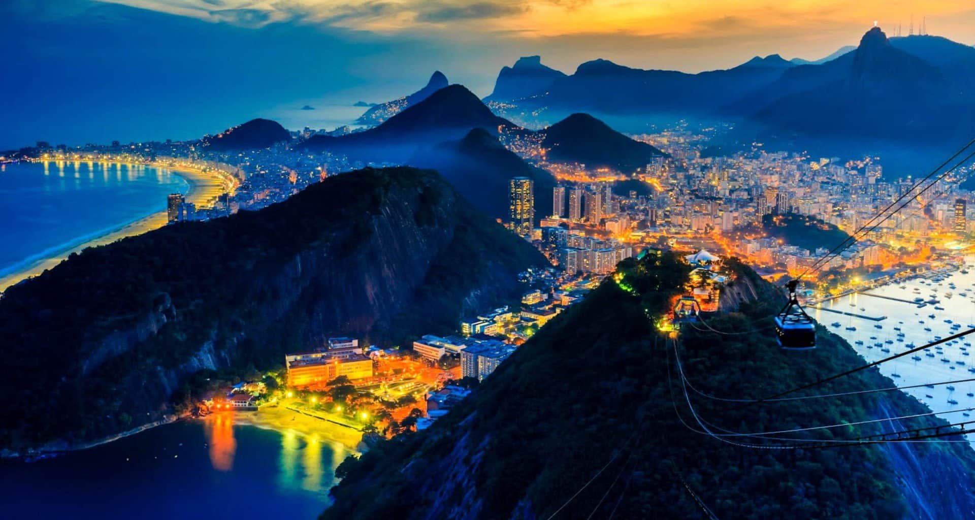 Aerial view of Rio de Janeiro at dusk, with city lights glowing, mountains and Sugarloaf Mountain in the foreground—a stunning highlight of any deluxe tour or South America travel adventure under a dramatic sky.