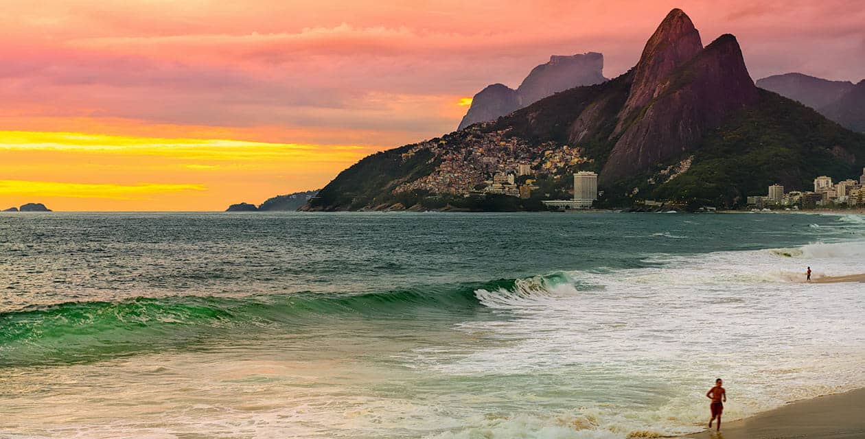 A person jogs along sandy beaches at sunset, with colorful skies, gentle waves, and lush mountains in the background near a coastal city.