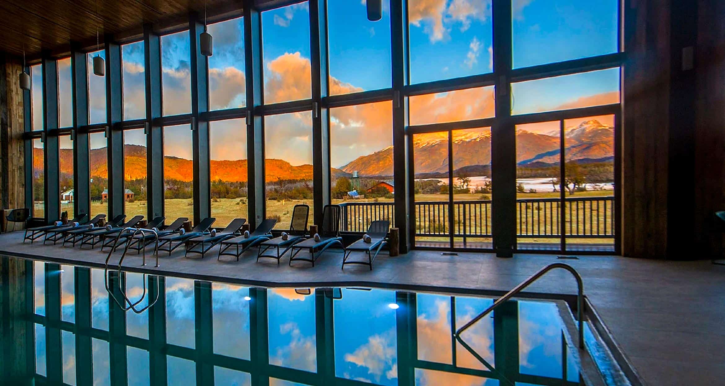 Indoor pool with lounge chairs beside large floor-to-ceiling windows at Rio Serrano Lodge, reflecting Torres del Paine's mountains, a Patagonian lake, and a dramatic sunset sky. Warm light illuminates the landscape and the pool’s calm water.