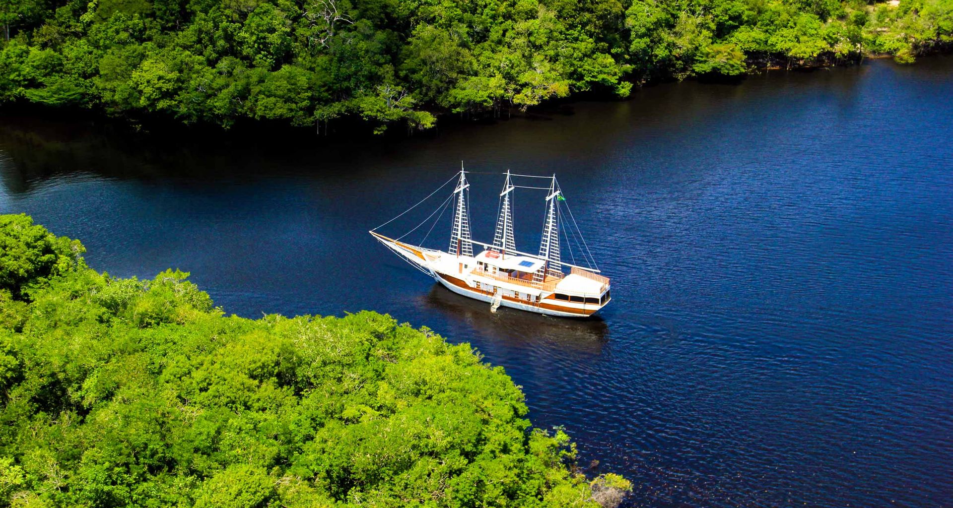 A white sailboat with three masts floats on a calm, dark blue river surrounded by dense, green forest—an enchanting scene reminiscent of a Brazil Amazon Cruise—viewed from above near the riverbank.