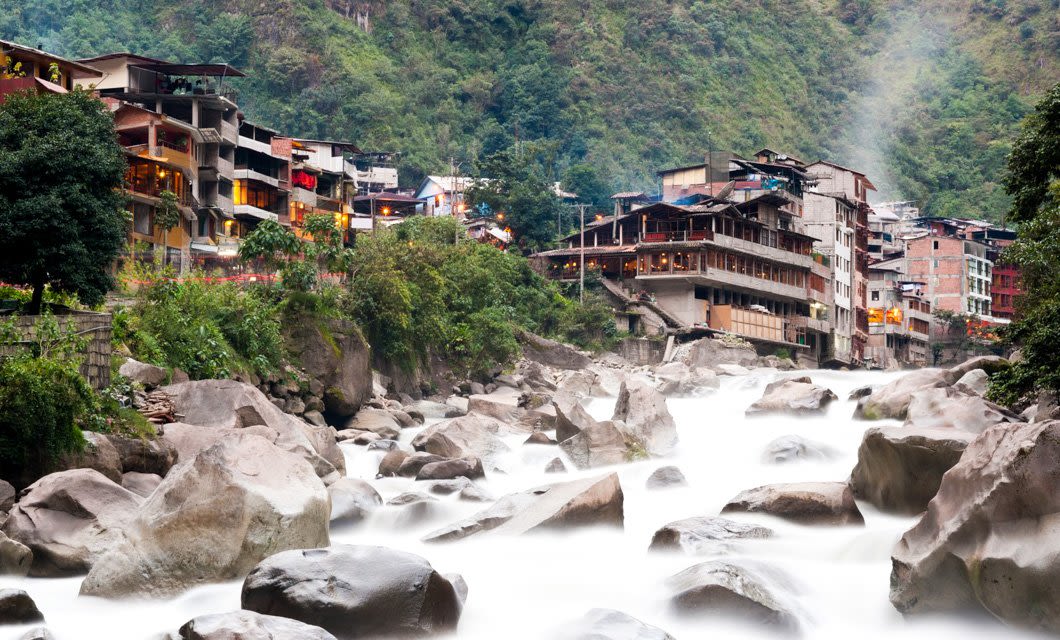 Buildings are perched on a lush, green hillside above a rocky river that winds through the mountains. Dense vegetation and mist surround the village, evoking the scenic landscapes found along the Salkantay Trek.