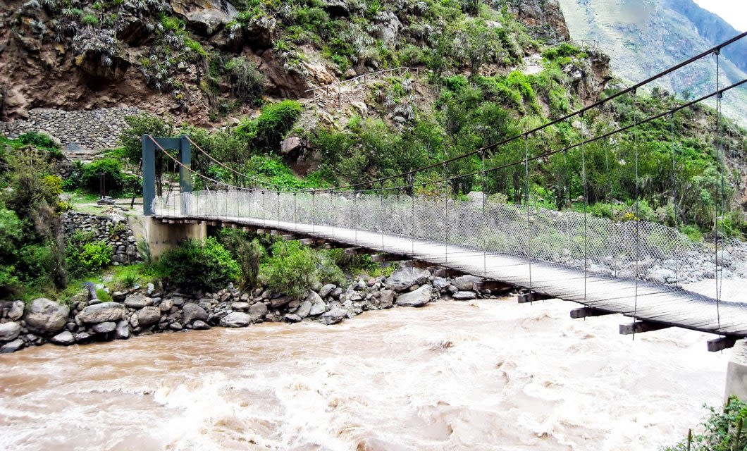 A narrow suspension bridge with metal railings crosses a fast-flowing, muddy river surrounded by rocks and lush green vegetation along the scenic Inca Trail to Machu Picchu in a mountainous area.
