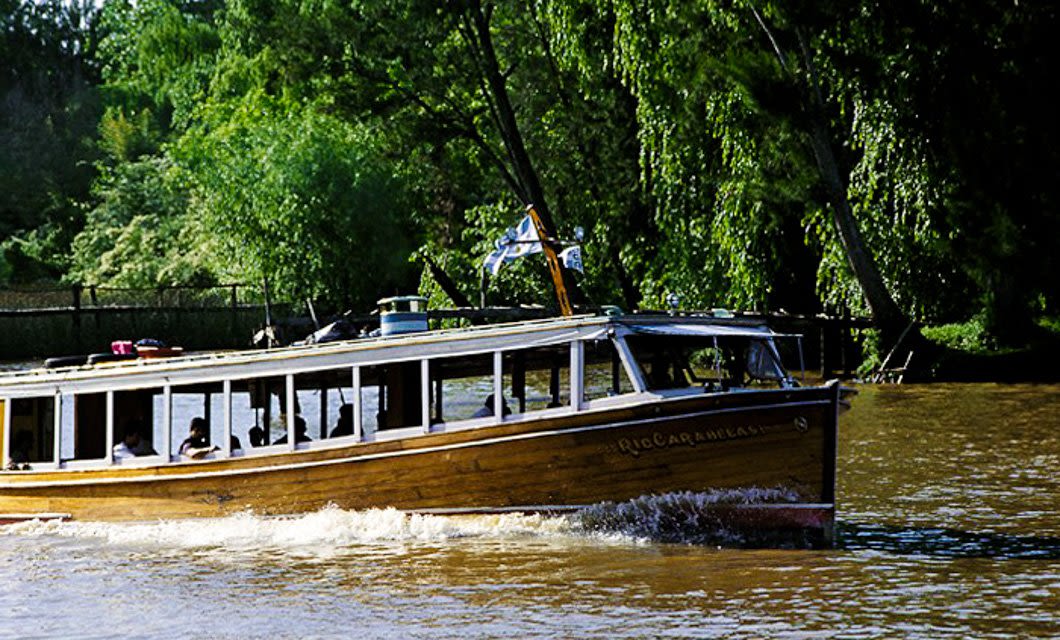 A wooden riverboat with windows and a blue-and-white flag glides through brown water near Buenos Aires, surrounded by lush green trees on a sunny day.