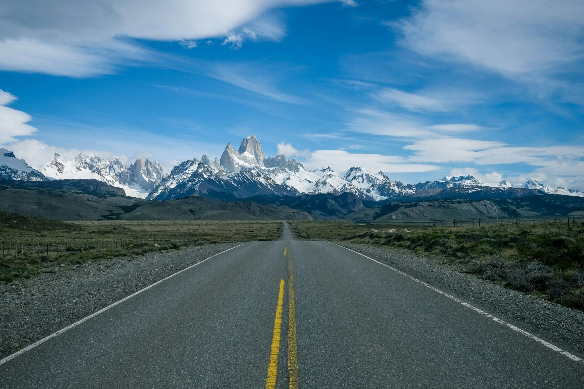 A straight, empty road leads toward distant snow-capped mountains under a bright blue Patagonian sky with scattered clouds. Grassy plains border the road, creating a dramatic landscape perfect for a guided group tour of Patagonia essentials.
