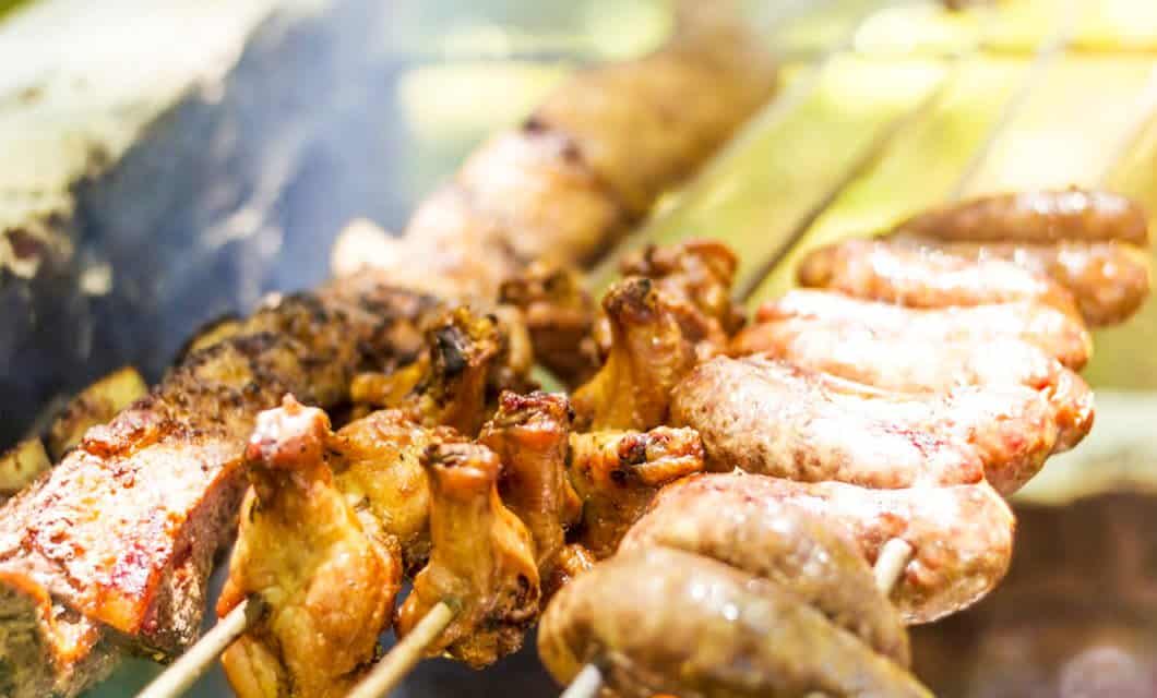 Close-up of assorted meats, including sausages, chicken, and ribs, cooking on skewers over a barbecue grill. The food appears juicy and golden-brown with a smoky background reminiscent of a feast near Iguazu Falls.