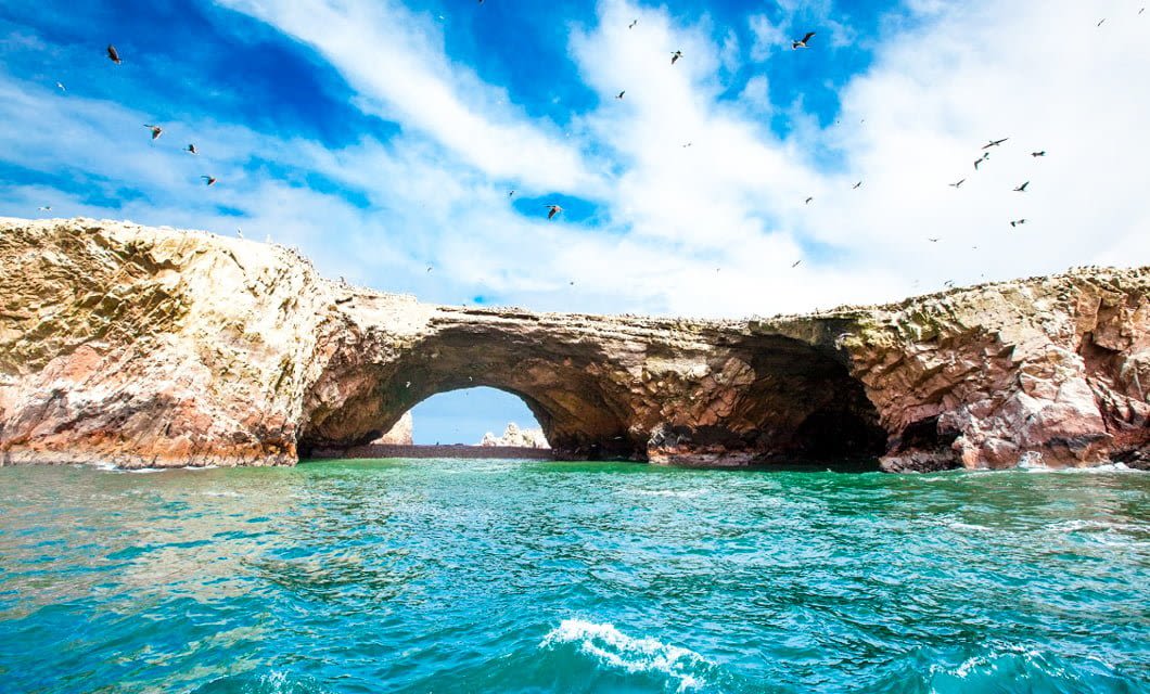Rocky coastal arch formation rises above turquoise water under a sunny blue sky in Peru, with seabirds flying overhead and waves gently lapping against the stone—capturing the essence of Peru’s dramatic shoreline.