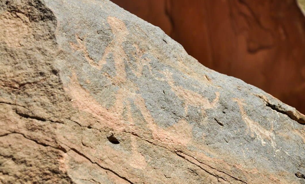 Ancient rock art in the Andes depicts a human figure standing beside two animals, possibly goats or deer, carved into a rough stone surface with a reddish background near Velasco.