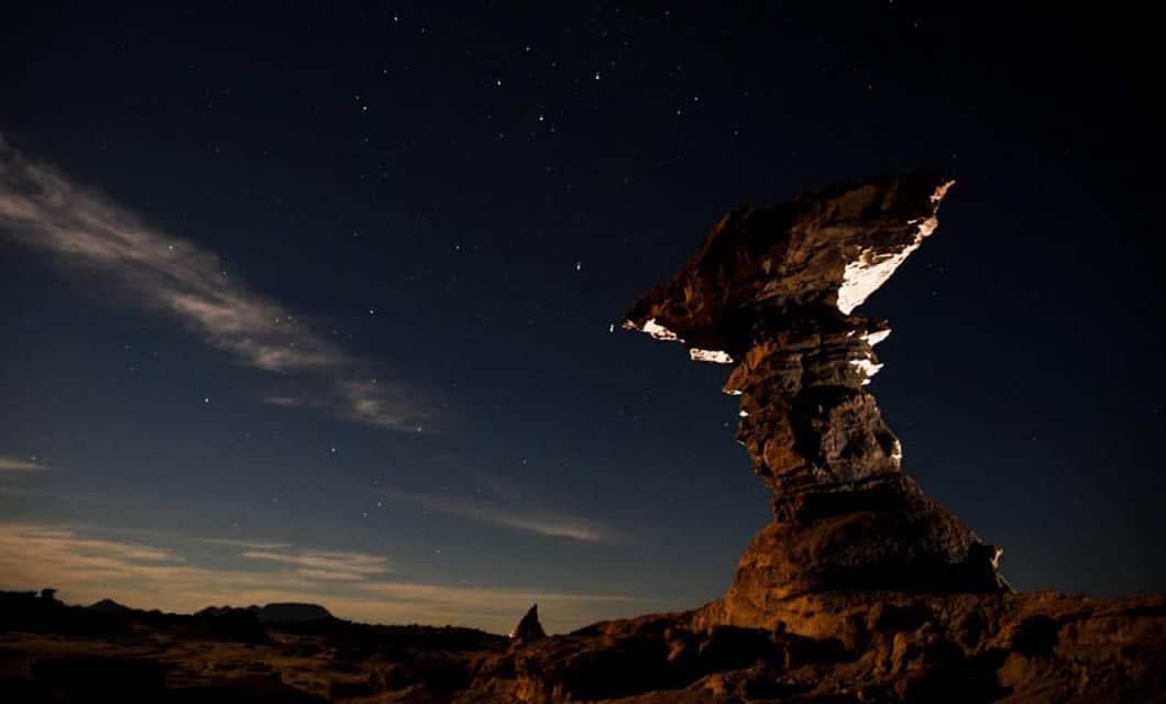 A large, uniquely-shaped Velasco rock formation stands silhouetted against a star-filled Andes night sky, with faint clouds on the left and subtle light illuminating parts of the rock.