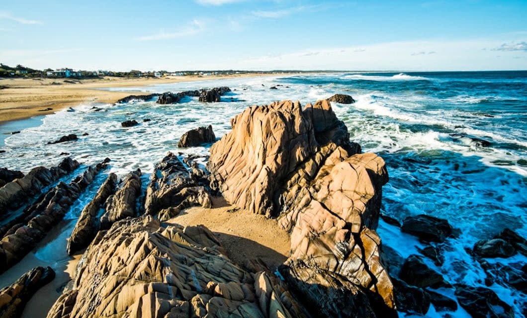 Rocky formations extend into the ocean, with waves crashing against them. A sandy beach stretches into the distance on the left, under a blue sky with scattered clouds—perfect for horseback riding along Uruguay’s picturesque coastline. Coastal houses are visible far in the background.