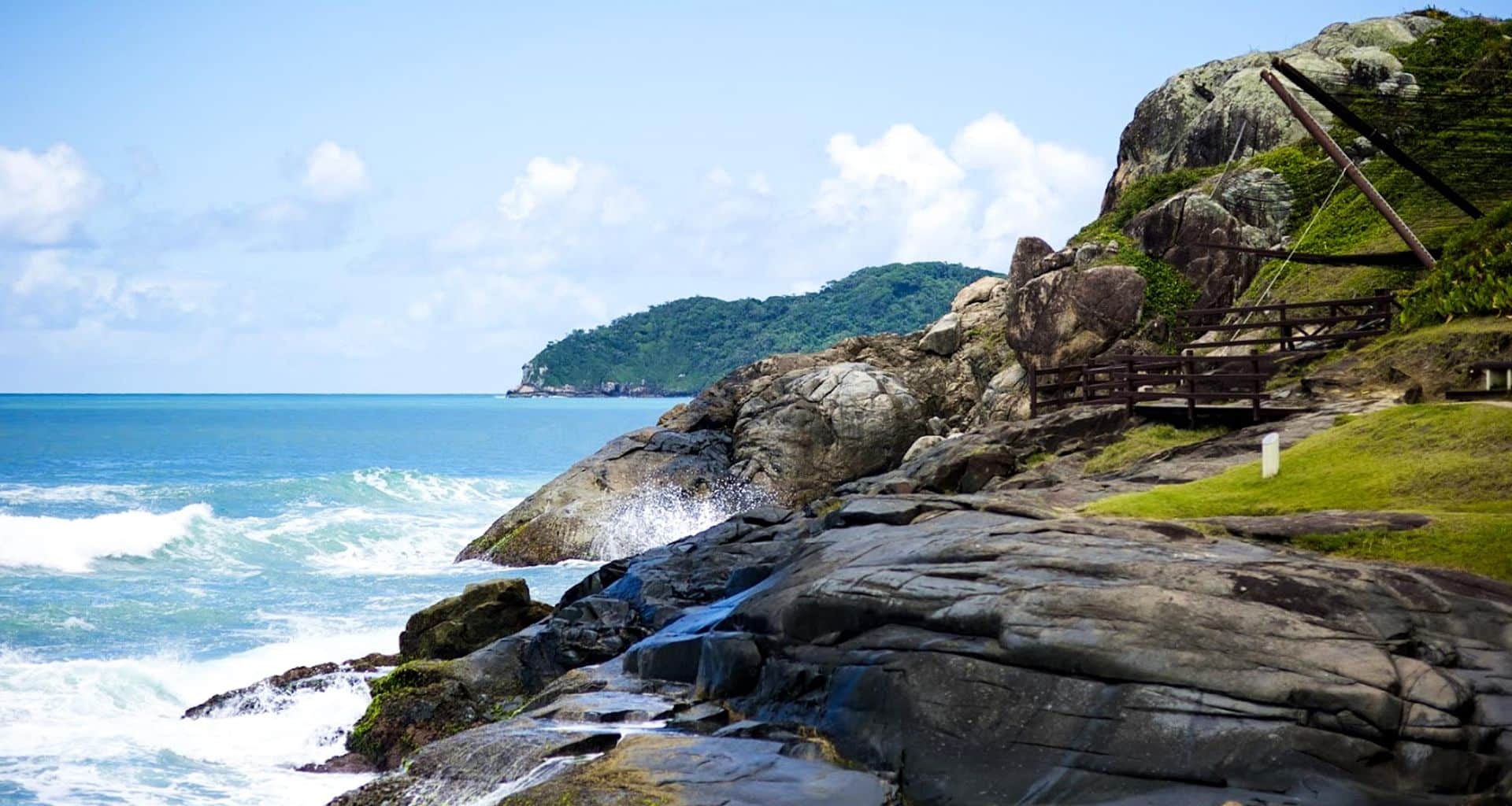 Rocky coastline with waves crashing against the rocks, a wooden fence runs along the edge, and lush green hills and blue ocean visible in the background under a partly cloudy sky on this Brazil Adventure Tour in Florianopolis, Santa Catarina.
