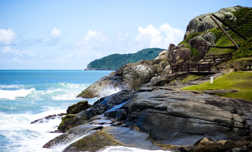 Rocky coastline with waves crashing against large rocks, a wooden fence running along a grassy area—perfect for adventure seekers in Southern Brazil—green hillside in the background, and blue sky with scattered clouds.