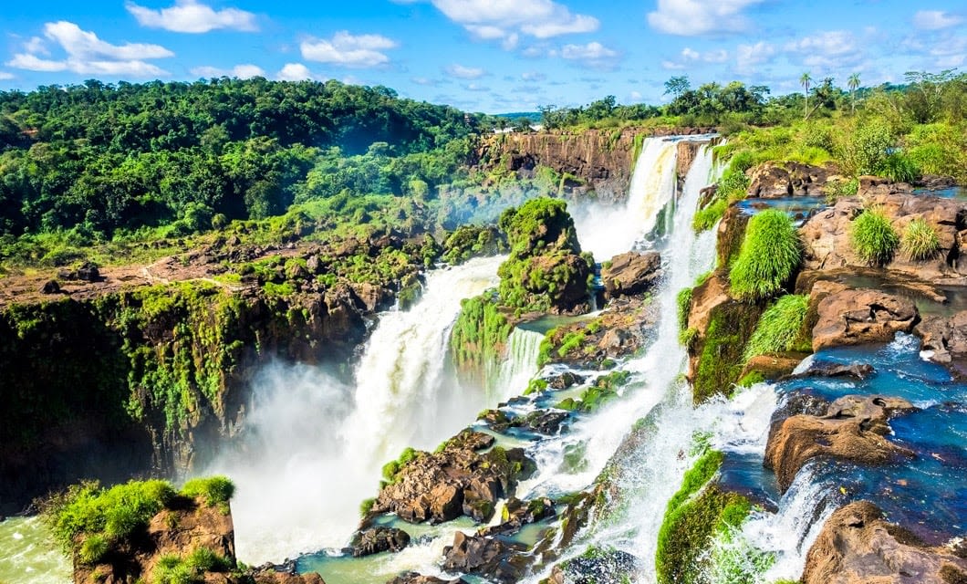 A scenic view of Iguazu Falls with powerful waterfalls cascading over rocky cliffs surrounded by lush green vegetation, reminiscent of landscapes once admired by the Incas, under a blue sky dotted with clouds.