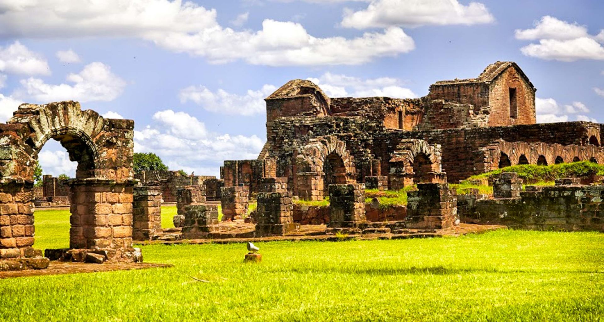 Ancient stone ruins of a Jesuit Missions complex stand on a vibrant green lawn under a blue sky with scattered clouds. The architecture features arches and weathered brick walls, reminiscent of sites near Iguazú Falls.