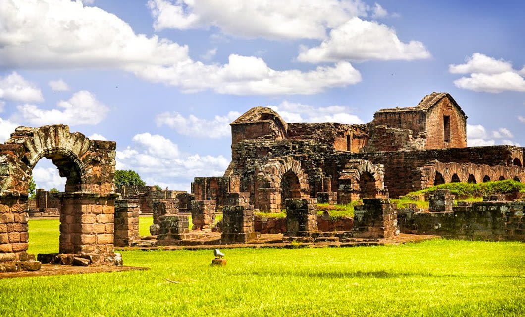 Ancient stone ruins of a Jesuit Missions complex with arches and partially standing walls, surrounded by green grass under a bright blue sky with scattered clouds, not far from the wonder of Iguazú Falls.
