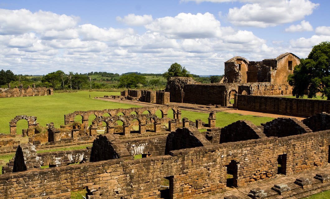 Ancient stone ruins of a mission complex stand under a partly cloudy sky near Asuncion, surrounded by green grass and trees, with arched doorways and weathered walls visible throughout the site.
