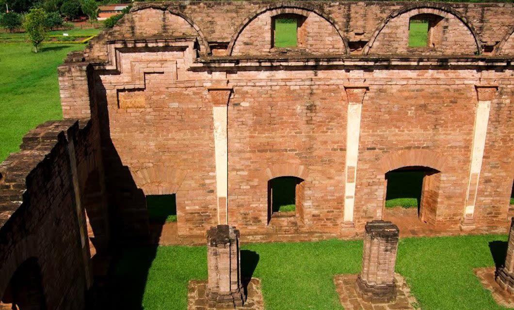 Ruins of an old brick building with arched windows and doorways, partially open to the green grass outside under bright sunlight—echoing the historic charm found near Asuncion and the wild beauty of Chaco.