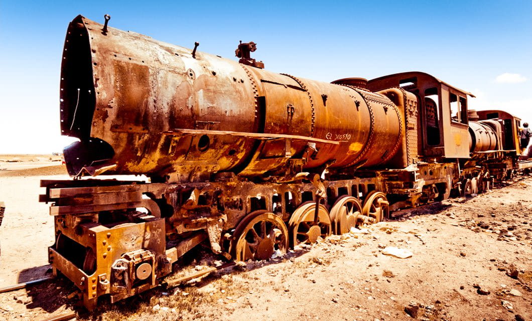 A rusted, abandoned steam locomotive sits on dry, barren ground under a bright blue sky—an iconic sight often featured on a Classic Bolivia Tour—with missing parts and extensive corrosion visible on its metal surface.