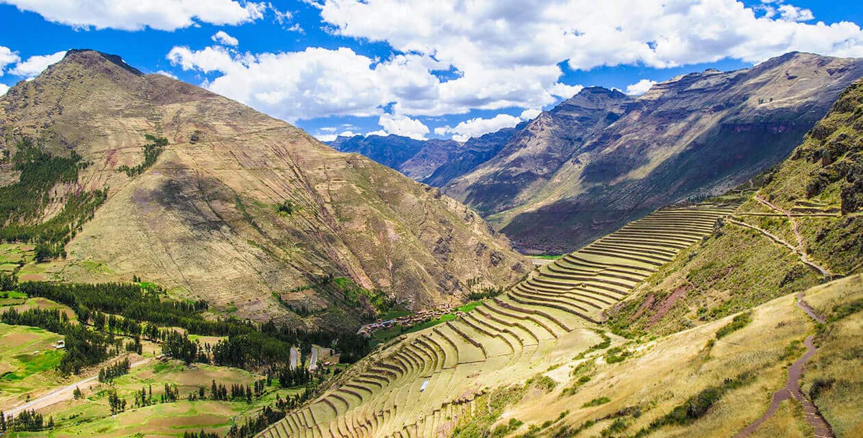 Terraced agricultural fields on a mountain slope in the Sacred Valley, Peru, showcase one of South America's natural wonders, with green valleys, towering Andean mountains, and a bright, partly cloudy sky.