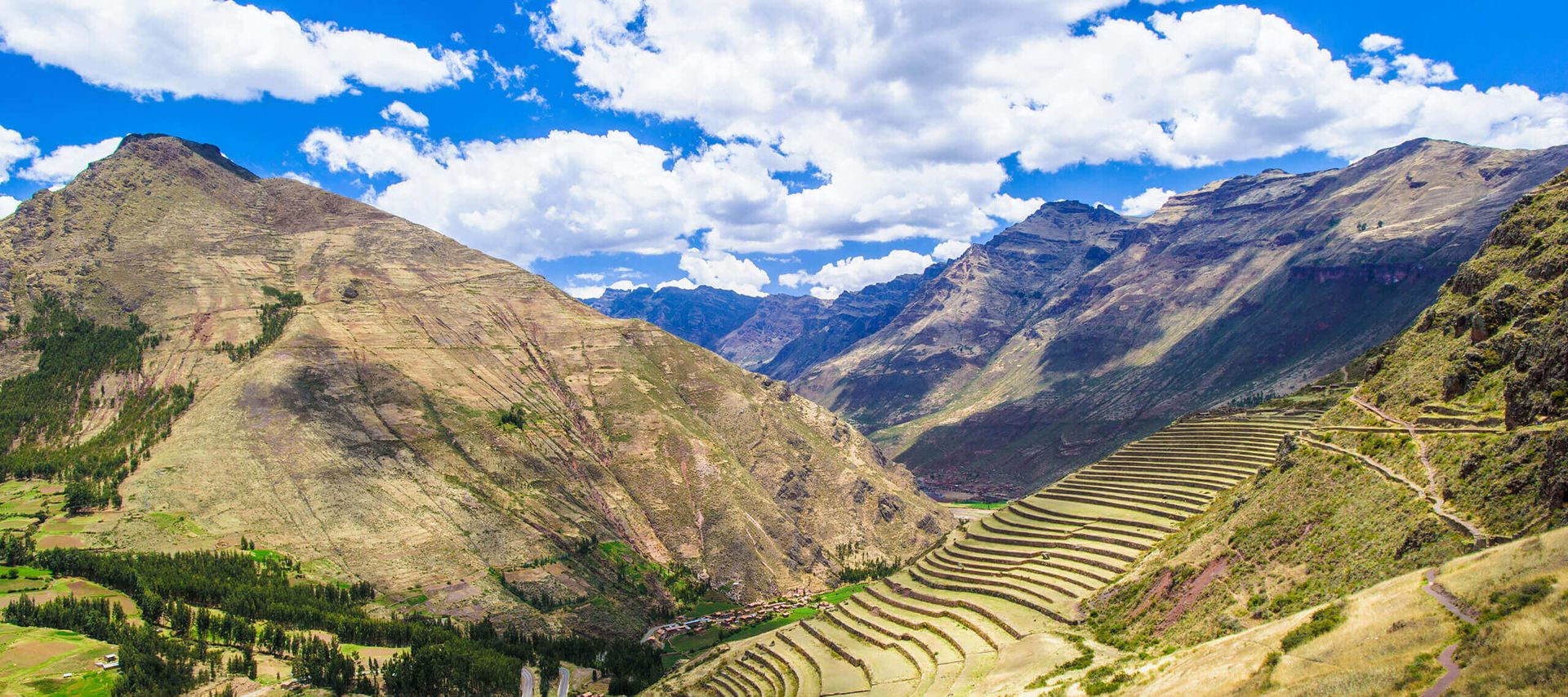 Panoramic view of green terraced fields on a mountain slope in South America, perfect for trekking, surrounded by towering mountains under a bright blue sky with scattered clouds.