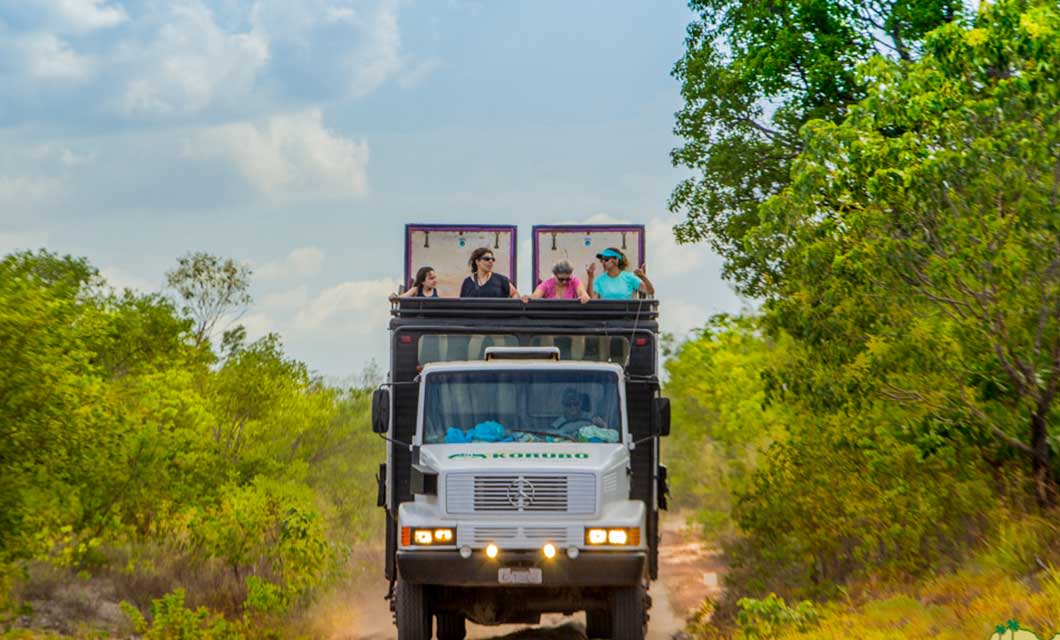 A safari truck with several people on its open top drives along a dirt path through Brazil's Cerrado in Jalapão, surrounded by lush green trees and bushes under a partly cloudy sky.