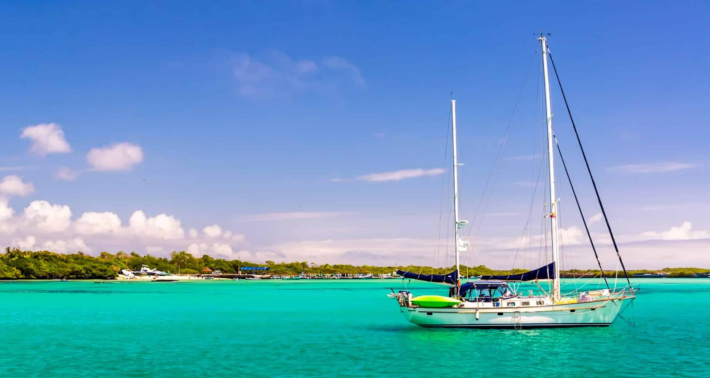 A sailboat floats on calm, turquoise water near a green shoreline under a bright blue sky with scattered white clouds in the Galapagos Islands. Other boats are visible near the shore in the background.