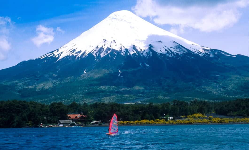 A windsurfer with a red sail glides across a blue Patagonia Lake, with a large snow-capped volcano and green forest in the background under a partly cloudy sky.