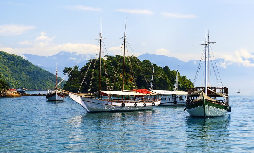 Traditional wooden boats anchored on calm blue water near a lush green island, with mountains and a partly cloudy sky in the background—perfect for exploring Colonial Gems on your Brazil Tour.