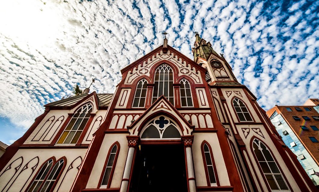 A Gothic-style church with tall arched windows and a pointed steeple stands under a dramatic sky, reminiscent of a tour through La Paz, with sunlight shining from the upper left corner and scattered clouds above.