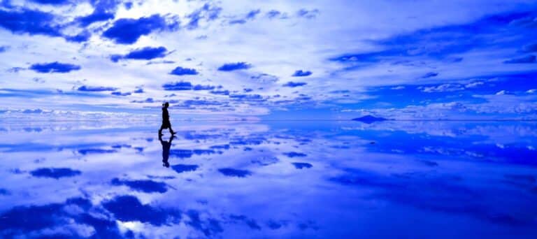 A person walks alone across a vast, reflective surface under a blue sky with scattered clouds, creating a mirror-like effect below—a dreamlike scene often experienced on Bolivia tours.
