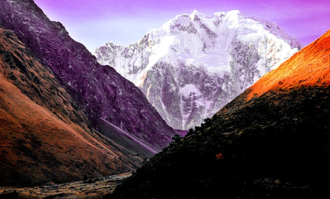 Snow-covered mountain peak under a purple sky, framed by steep, shadowed hillsides with one side bathed in golden sunlight and a narrow valley—an iconic view along the Salkantay Trek near Salkantay Lodge.