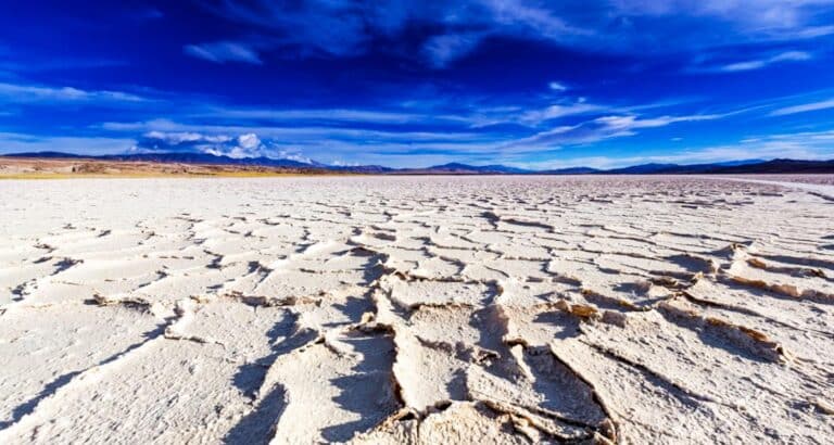 A wide expanse of cracked, dry earth under a vibrant blue sky with scattered clouds, surrounded by distant mountains. The scene depicts an arid desert landscape reminiscent of the striking vistas near Cafayate in Salta.