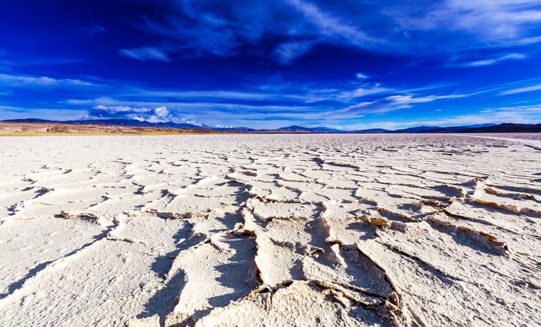 Cracked dry land stretches across a barren landscape under a vibrant blue sky with wispy clouds, distant mountains on the horizon, evoking the arid beauty of Cafayate and Purmamarca in Salta.