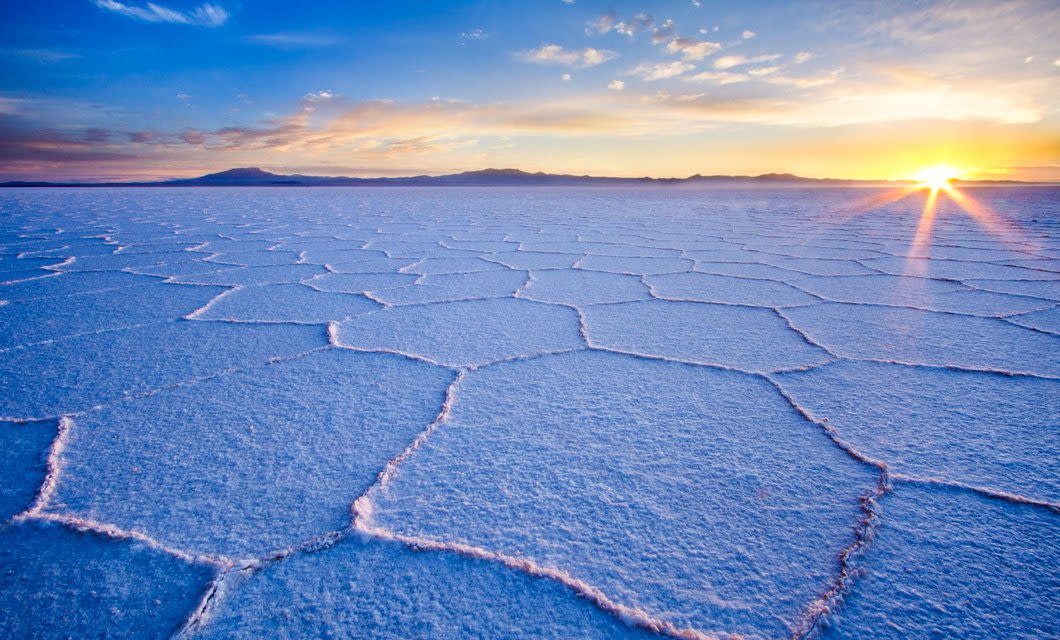 Sunrise over the vast, geometric salt flats of Bolivia reveals a stunning pattern of hexagonal shapes, under a bright sky with scattered clouds and distant mountains on the horizon.