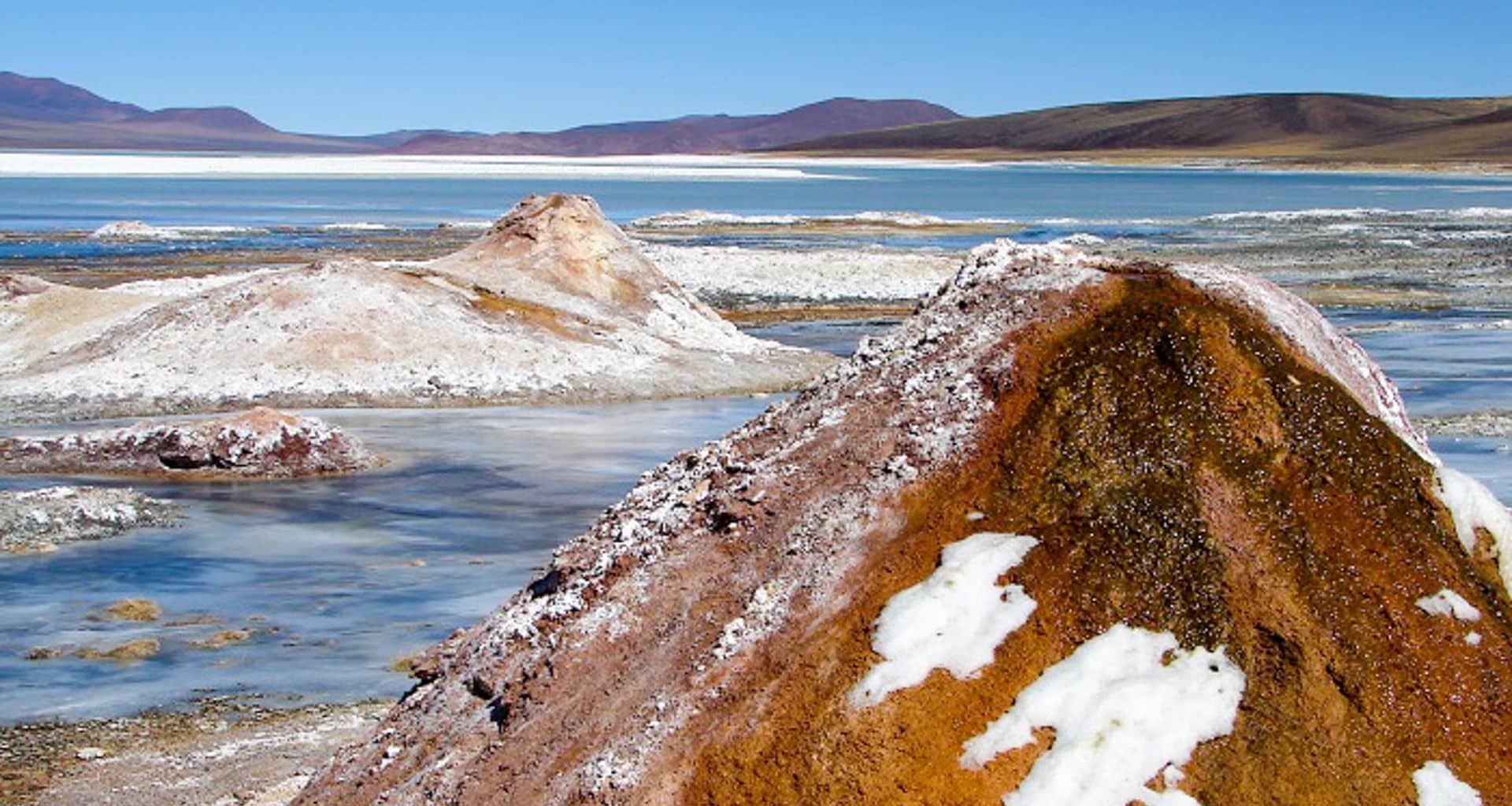 Salt mounds with patches of white crystals and mineral deposits beside a shallow blue lake, surrounded by the arid, brown hills and mountains of the Andes near Velasco, all under a clear blue sky.