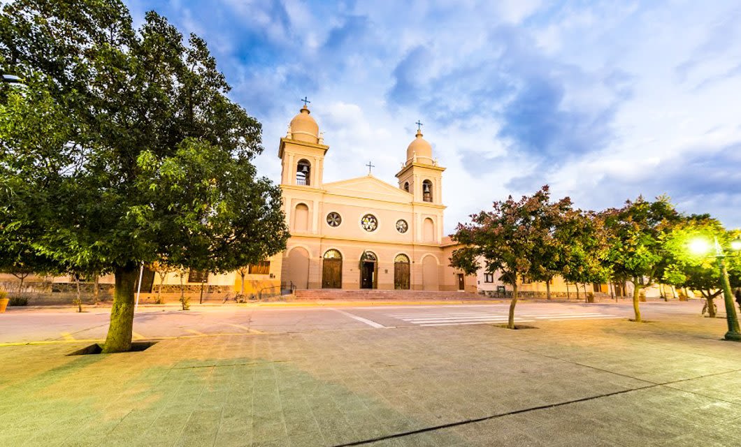 A large, historic church with two bell towers and arched windows stands in an empty plaza lined with trees under a cloudy Salta sky at dusk.