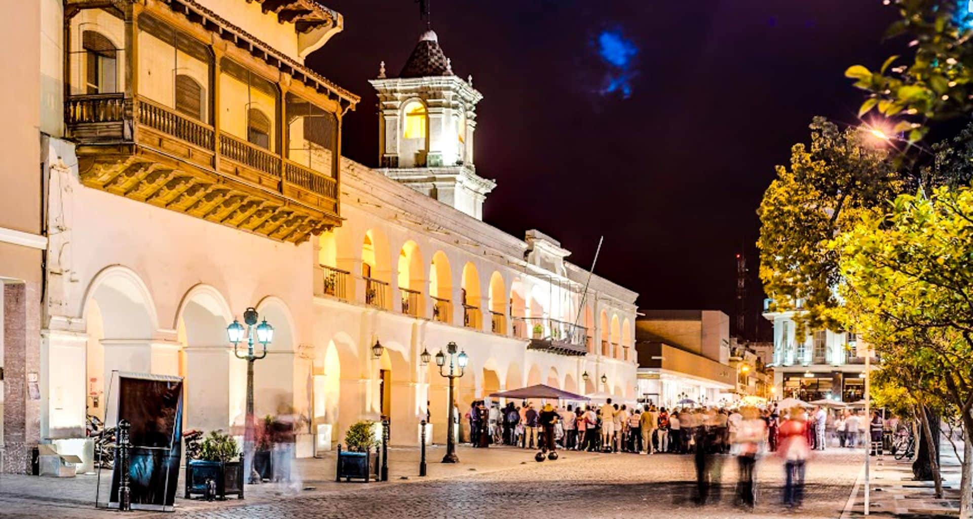 Night view of a brightly lit colonial-style building with arches and a balcony, bustling with people as if awaiting a railway journey in a lively plaza under a dark sky. Trees and lampposts line the street.